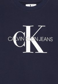 Námořnická modrá bavlněná mikina s kulatým výstřihem; na prsou se nachází výrazný bílý text "CALVIN KLEIN JEANS" umístěný ve středu.