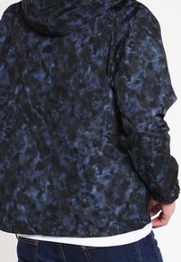 Veste à capuche en bleu foncé et noir, avec un motif abstrait texturé. Matériau léger avec des poignets élastiques et une coupe décontractée.