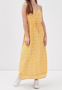 Robe maxi en imprimé floral jaune avec des bretelles spaghetti ajustables et une taille froncée. Des baskets blanches complètent le style décontracté.