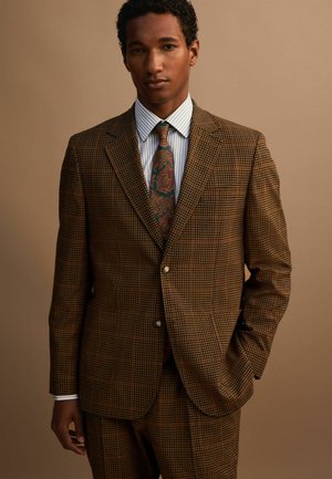 Man wearing a brown checkered suit with a striped white shirt and a paisley patterned tie, standing against a beige background.