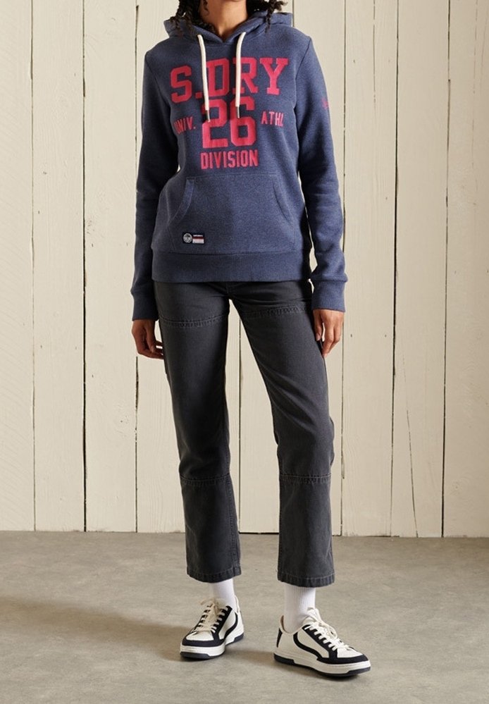 Personne portant un sweat à capuche bleu avec des lettres rouges, un pantalon court noir, des chaussettes blanches et des baskets noir et blanc, debout sur un sol gris.