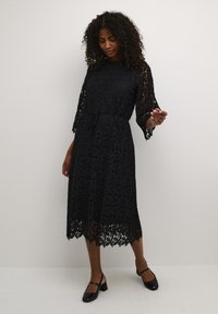 Black lace dress with floral pattern, three-quarter sleeves, cinched waist, mid-length, and scalloped hem. Worn with black shoes.