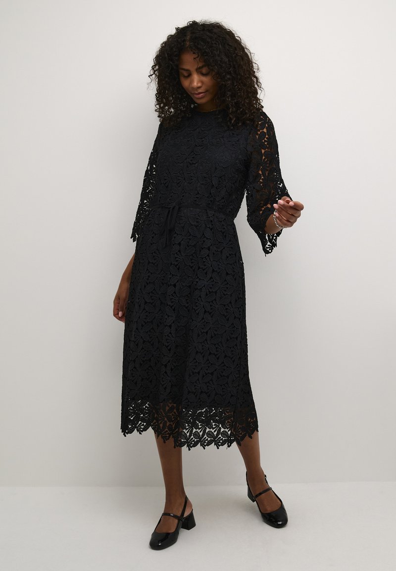 Black lace dress with floral pattern, three-quarter sleeves, cinched waist, mid-length, and scalloped hem. Worn with black shoes.