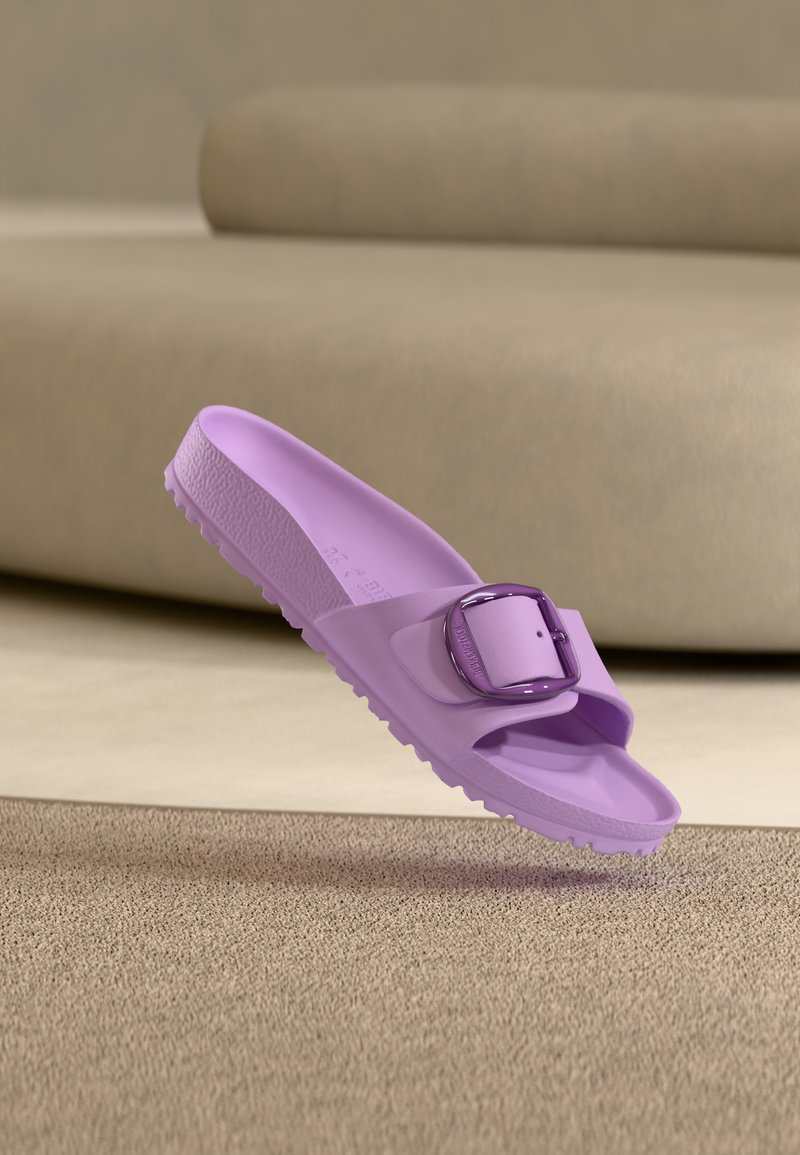 Μωβ σανδάλι με φαρδιά αγκράφα πάνω σε μπεζ χαλί μπροστά από μια μπεζ καναπέ.