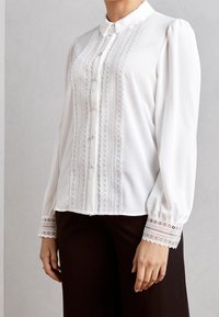 Blouse blanche avec col ajusté, détails en dentelle verticale et fermeture par boutons. Comprend des manches bouffantes et des poignets en dentelle.