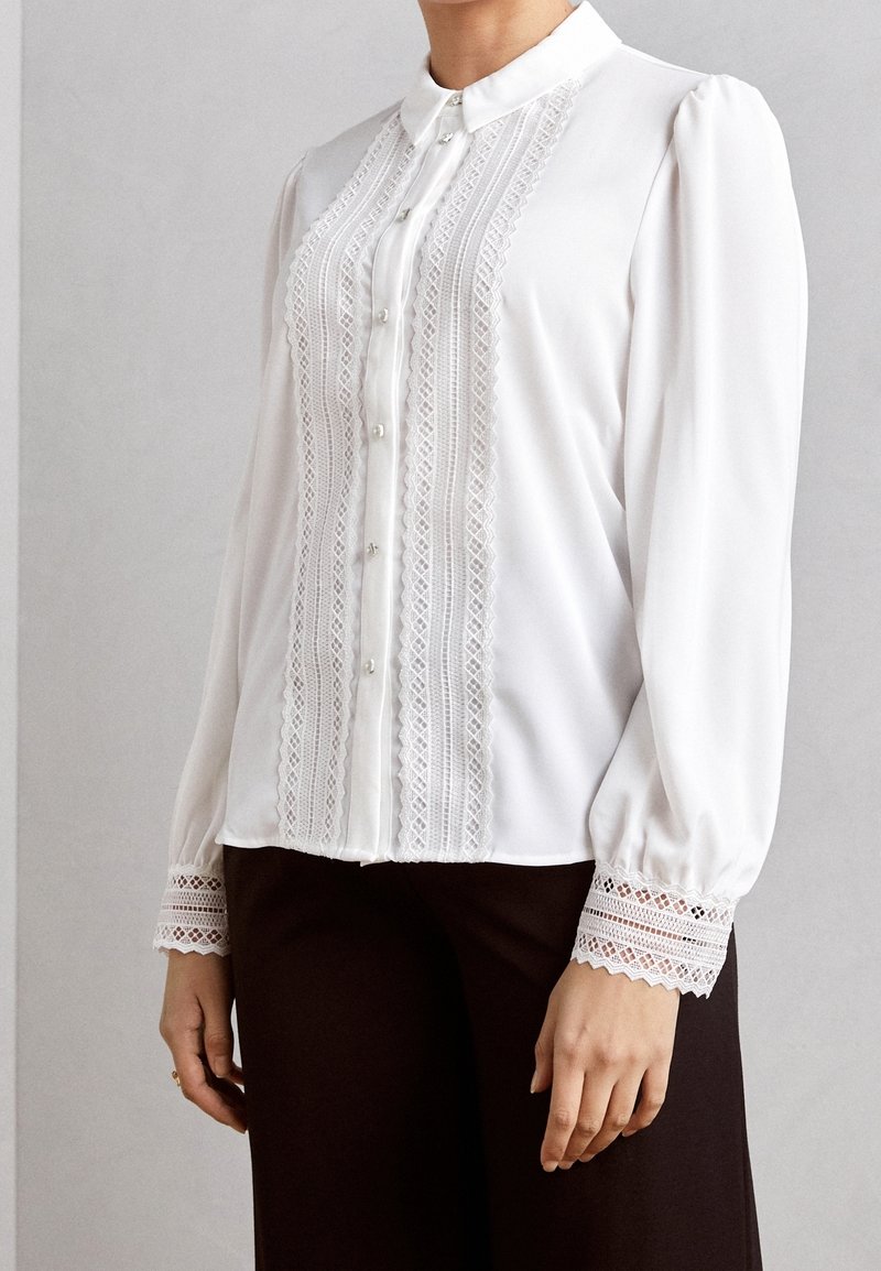 Blouse blanche avec col ajusté, détails en dentelle verticale et fermeture par boutons. Comprend des manches bouffantes et des poignets en dentelle.