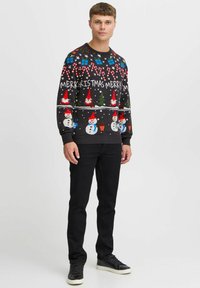 Suéter negro con coloridos diseños navideños, incluyendo muñecos de nieve, bastones de caramelos, árboles y texto, combinado con pantalones negros y zapatillas.