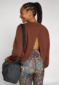 Top long à manches marron avec un design à dos ouvert, associé à un legging à motifs multicolores et un sac à épaule noir avec zip.