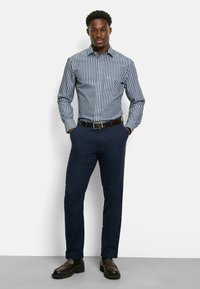 Camicia a righe blu navy e bianca, abbinata a pantaloni blu navy scuri. Il modello è in piedi con le mani in tasca, indossando scarpe marroni.