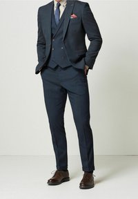 Granatowy garnitur składający się z trzech części, wykonany z teksturowanej tkaniny, z klapami w kształcie notched, kamizelką oraz brązowymi skórzanymi butami.