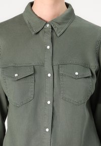 Camisa de botão verde-oliva feita de tecido suave, com colarinho pontiagudo, dois bolsos no peito e botões de pressão prateados.