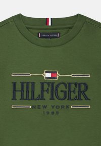Πράσινο βαμβακερό μπλουζάκι με κεντημένο σε σκούρο μπλε το κείμενο "HILFIGER" και μικρό κόκκινο, λευκό και μπλε διακοσμητικό λογότυπο στη λαιμόκοψη.