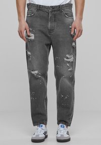 Jeans de mezclilla negra con desgastes, áreas descoloridas y salpicaduras de pintura blanca. Diseñados con un corte holgado y estilo clásico de cinco bolsillos.
