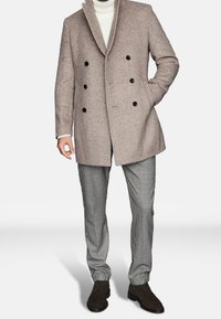 Cappotto di lana a spina di pesce marrone chiaro, doppiopetto, con bottoni neri. Abbinato a pantaloni a quadri bianchi e neri e scarpe marroni.