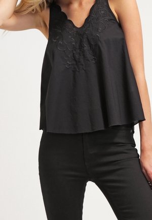 Vrouw draagt een mouwloos zwart topje met bloemenborduursel bij de halslijn en strakke zwarte broek, poseert met opgetaste arm.