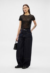 Νέα γυναίκα με τατουάζ φορά διάφανο μαύρο τοπ με κείμενο, φαρδιά σκουρόχρωμα τζιν, μαύρα παπούτσια και κρατά μικρή μαύρη τσάντα, στέκεται όρθια.