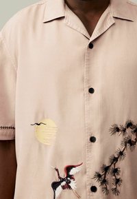 Beżowa koszula z krótkim rękawem i czarnymi guzikami. Posiada haftowanego żurawia oraz słońce, z misternym wzorem gałązki sosny na tkaninie.
