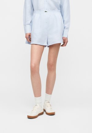 Femme portant un short bleu clair et une chemise assortie, des chaussettes blanches et des baskets blanches à semelles marron, debout sur un fond uni.