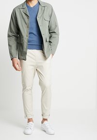 Jasnozielona, krótka kurtka z zapięciem na guziki i wieloma kieszeniami, noszona na niebieskim swetrze z dekoltem w serek. W połączeniu z beżowymi spodniami taperowanymi i białymi trampkami.