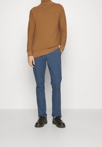 Homme portant un pull à col roulé marron texturé, un pantalon bleu slim, des chaussures noires à lacets, debout avec une main dans la poche contre un mur blanc.