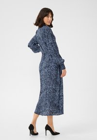 Robe bleu marine à manches longues avec un motif abstrait blanc, taille cintrée, jupe fluide et longueur totale. Stylée avec des chaussures à talons hauts noires.