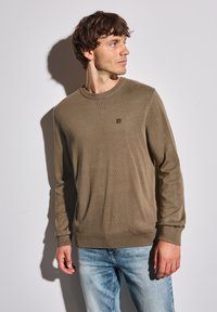 Khaki crewnecktröja med långa ärmar, stickat tyg, ribbade mudd och avslut, med en liten logotyp på framsidan. Bärs med blekta blå jeans.
