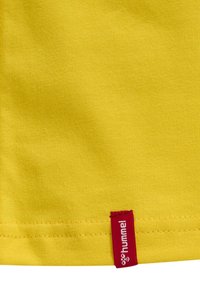Tessuto di cotone giallo con una texture liscia. Presenta un'etichetta rossa con il logo "hummel" sul bordo inferiore. Design pulito e semplice.