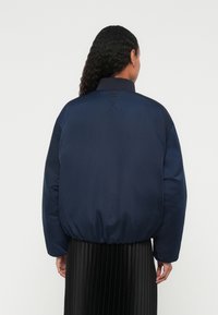 Navy blue puffer jacket with a ribbed collar, elastic waistband, and a smooth texture. No visible logos or patterns. Model wearing a pleated skirt.