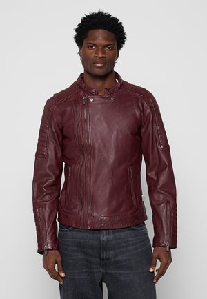 Mann steht frontal, trägt eine burgunderrote Leder-Bikerjacke mit asymmetrischem Reißverschluss und dunkelgraue Jeans vor einem schlichten Hintergrund.