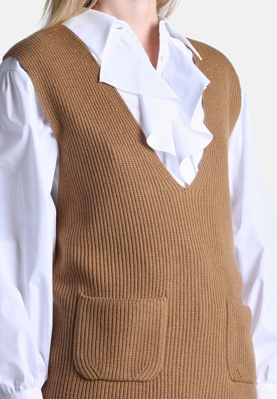 Blouse blanche avec grand col à volants sous un gilet sans manches marron en maille avec un col en V profond et deux poches avant.