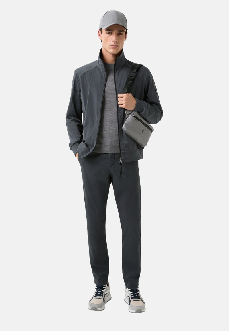 Dunkelgrau Herrenjacke mit hohem Kragen, graues T-Shirt, passende Hose und kleine graue Umhängetasche. Graue Kappe und Turnschuhe mit hellen Akzenten.