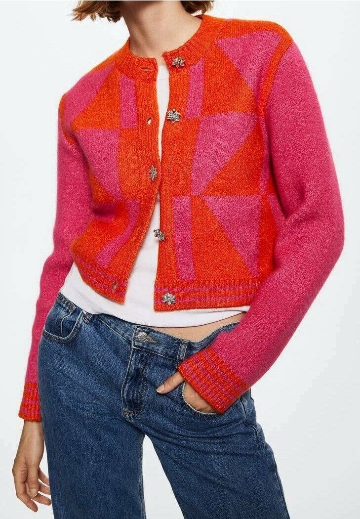 Femme portant un cardigan court à motif géométrique rose et orange avec des boutons décoratifs, un haut blanc et un jean bleu, main dans la poche.