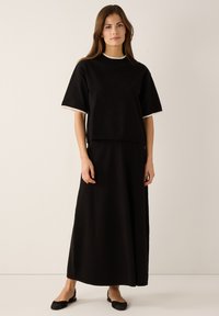 Haut noir à manches courtes avec col rond et bordure beige, associé à une longue jupe noire. Les deux pièces présentent une texture lisse et un design simple.