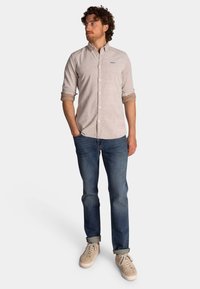 Lichtbeige overhemd met opgerolde mouwen, blauwe jeans en beige sneakers. Het overhemd heeft een klein logo op de borst.