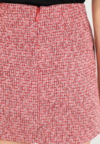 Mini-jupe en tweed rouge avec un motif texturé et une fermeture éclair rouge visible au dos. Tissu lisse avec une coupe structurée.