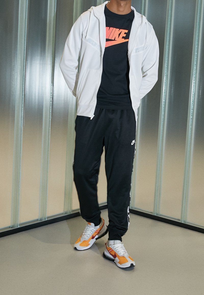 Biała bluza z zamkiem, czarny T-shirt z czerwonym logo Nike, czarne spodnie dresowe z bocznymi paskami oraz pomarańczowo-białe buty sportowe. Gładkie tekstury.