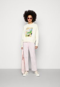 Sweatshirt creme com estampa gráfica, calças largas rosa, ténis brancos e óculos de sol rosa. Segurando um lenço leve com padrão rosa.