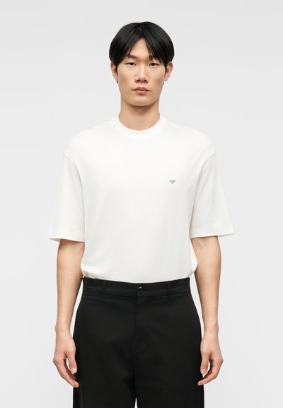 Άνδρας με ίσια μαύρα μαλλιά, φοράει απλό λευκό πουκάμισο με κοντά μανίκια και μαύρο παντελόνι, στέκεται μπροστά σε ανοιχτογκρι φόντο.