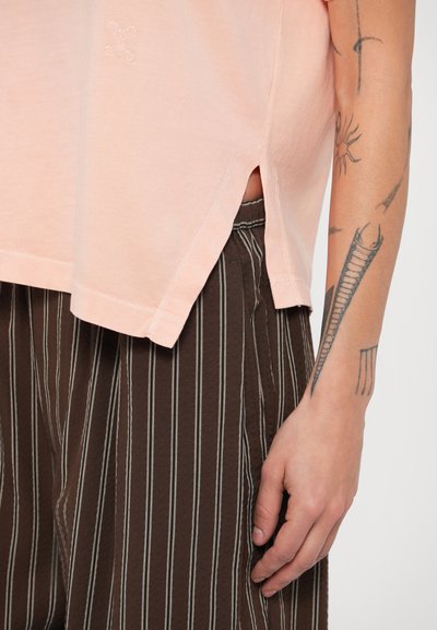 Ροζ βαμβακερό T-shirt με ανοίγματα στο πλάι και κεντημένο λογότυπο. Συνδυάζεται με καφέ ριγωτό παντελόνι, δείχνοντας χέρι με τατουάζ.