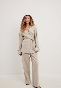 Bežova bluzica z vezanjem v pasu in zvonastimi rokavi, v kombinaciji s širokimi hlačami iste barve; obe imata teksturirano blago.