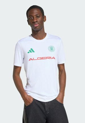 Ung mand med hænderne i lommerne iført hvid Algeriet fodboldtrøje med grønne og røde logoer, stående mod en ensartet lys baggrund.