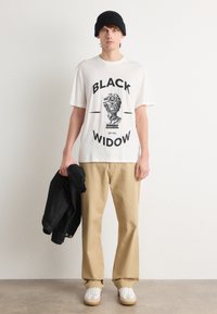 Hvit t-skjorte med "BLACK WIDOW" grafikk, beige bukser, svart lue, og denimjakke, med hvite sneakers med beige såler.