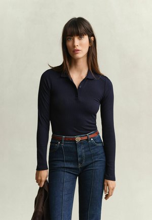 Femme aux longs cheveux bruns portant un haut à manches longues côtelé bleu marine et un jean bleu foncé, tenant une veste marron dans sa main gauche.