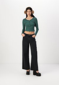 Vihreä uritettu pitkähihainen crop-top, jossa on napit, yhdistettynä leveälahkeisiin mustiin farkkuihin ja mustiin koroketossuihin. Leopardi-kuosiset korvalaput korostavat asua.