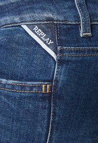 Mörkblå denimjeans med en bakficka som har en vit etikett med texten "REPLAY", förstärkt sömnad och en klassisk design.