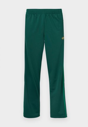 Pantalon de sport vert avec une ceinture élastique, présentant des bandes latérales et un petit logo doré sur le haut à gauche. Texture lisse.