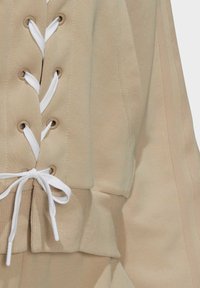 Béžová mikina se šněrováním vpředu, s bílými tkaničkami a kovovými průchody. Texturovaná tkanina a žebrované manžety.