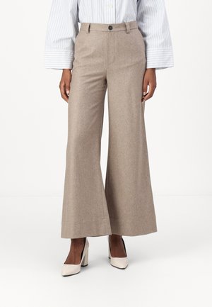 Pantalons à jambes larges de couleur marron clair en tissu texturé avec une fermeture à un bouton et des poches. Associés à des chaussures à talons hauts blanches.