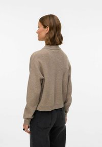 Bežs adīts džemperis ar apkakli, nolaižamām plecu šuvēm un saīsinātu apakšējo malu, kombinēts ar melniem augstās jostasvietas džinsiem. Gluda tekstūra, ikdienišķa piegriezuma stils.