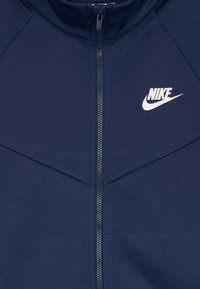 Tumši zila jaka ar augstu apkakli, centrālo rāvējslēdzēju un baltu Nike logotipu uz krūts; tam ir teksturēta, gluda auduma virsma.